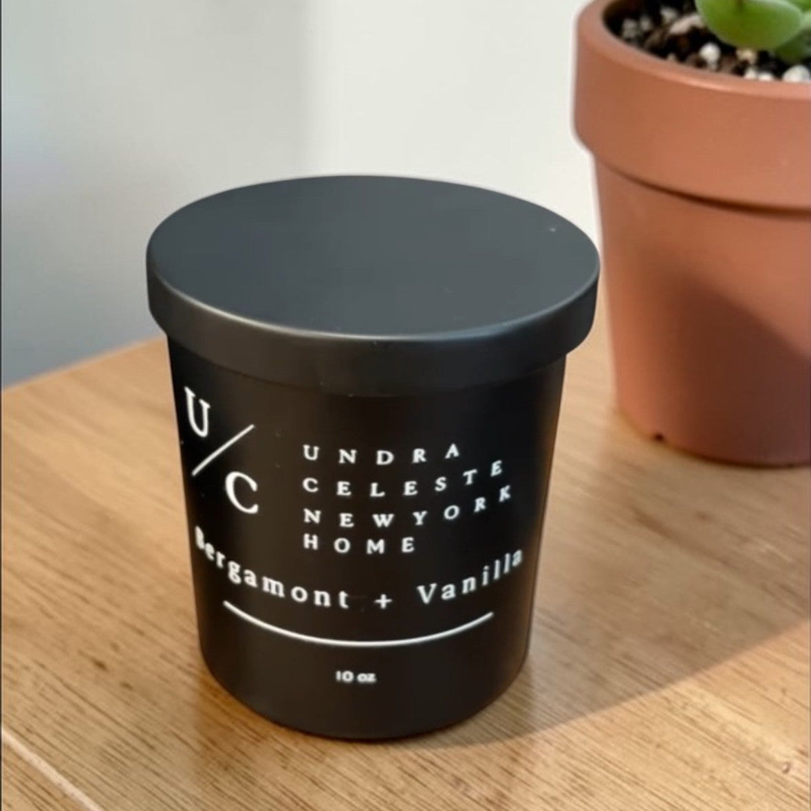 Black candle with white text on a wooden surface next to a potted plant. Bergamont and vanilla soy candle with wood wick.