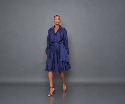 A model stands against a dark gray studio backdrop wearing a dark blue wrap-style midi dress with long sleeves and pleated skirt details, paired with tan heeled sandals.